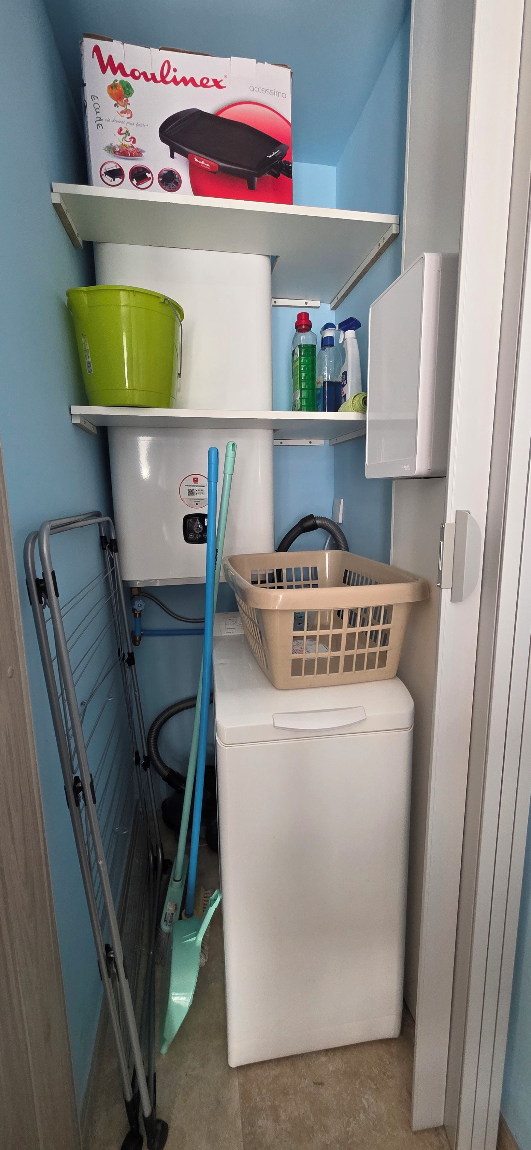 Un lavadero pequeño con lavadora, tendedero, un cubo verde y una cesta.