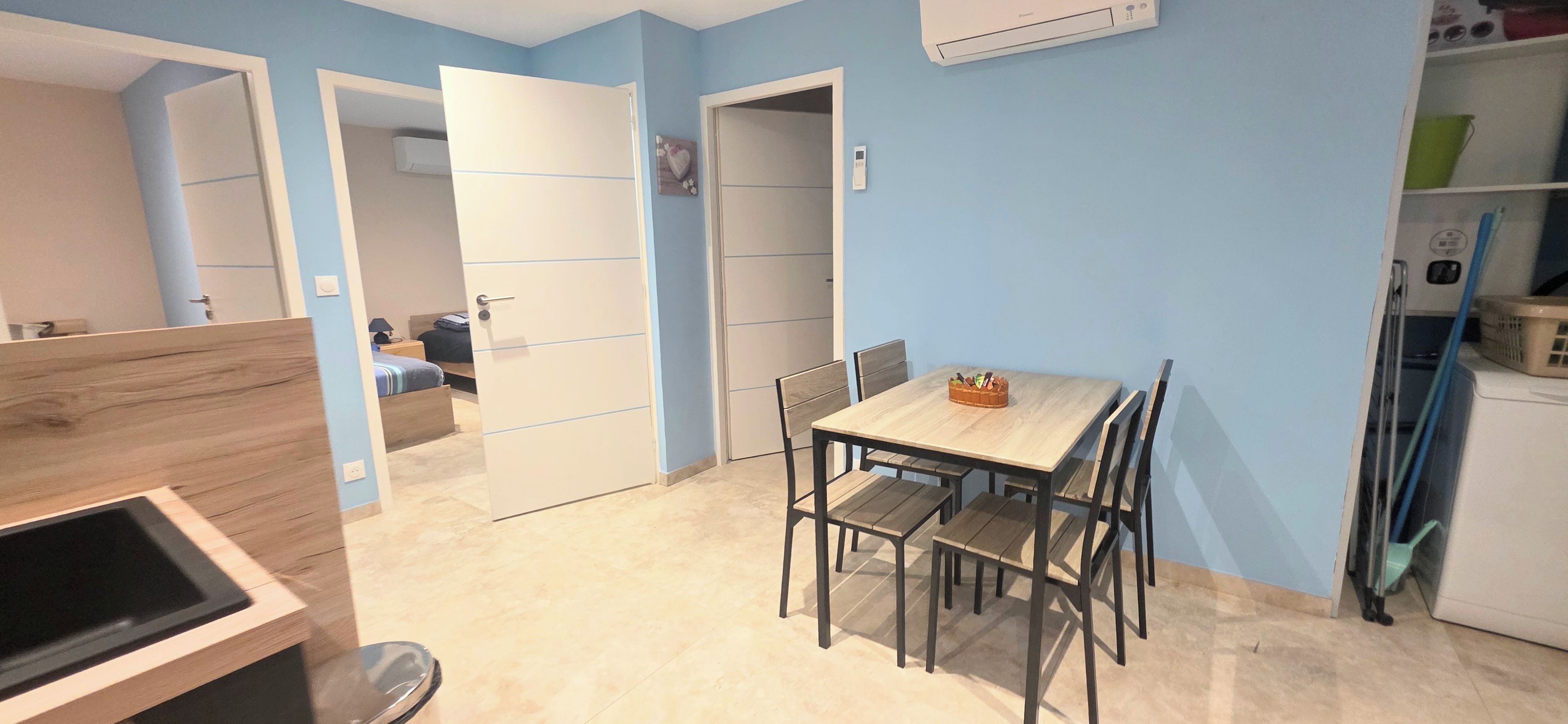 A compact kitchen with a dining table and chairs, a visible refrigerator, and a door leading to another room.