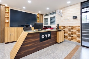 A reception desk with a dark wood finish, a glass top, and a sign that reads 'YO'. There are wooden cabinets to the left, a blackboard, and a window with blinds to the right.