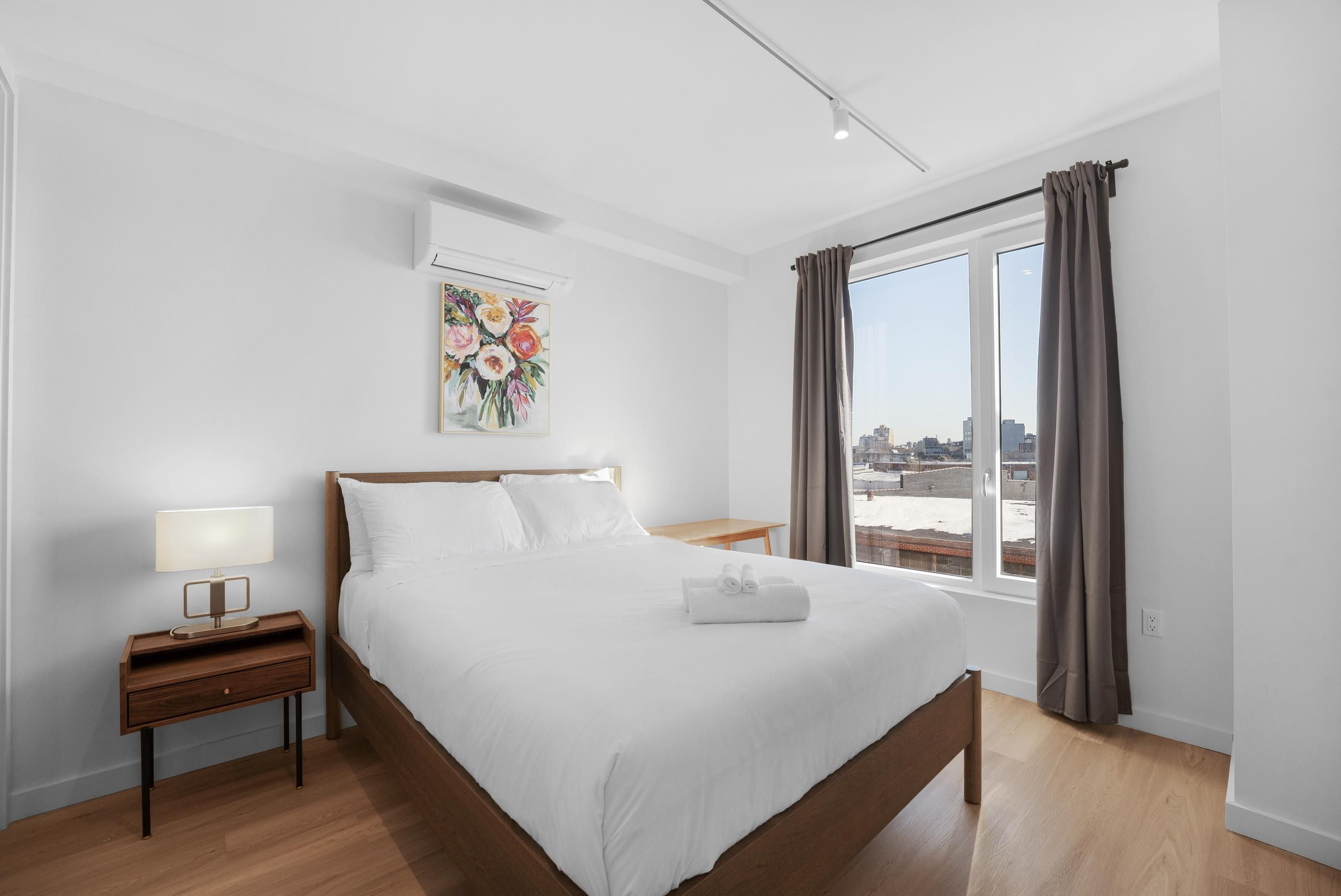 A hotel room with a large bed, a bedside table with a lamp, a painting on the wall, and a view of the city through the window.
