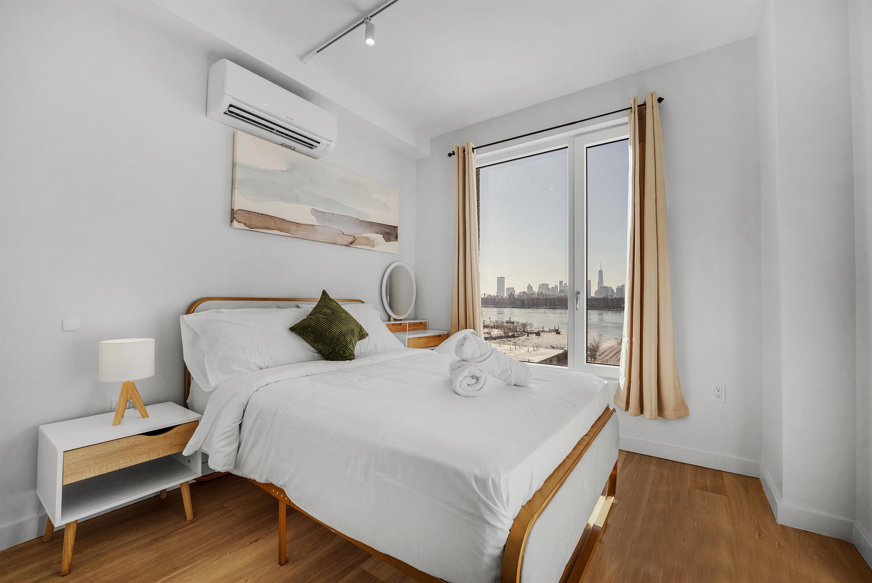 A modern hotel room with a large bed, a bedside table with a lamp, a view of a cityscape through a large window, and a wooden floor.