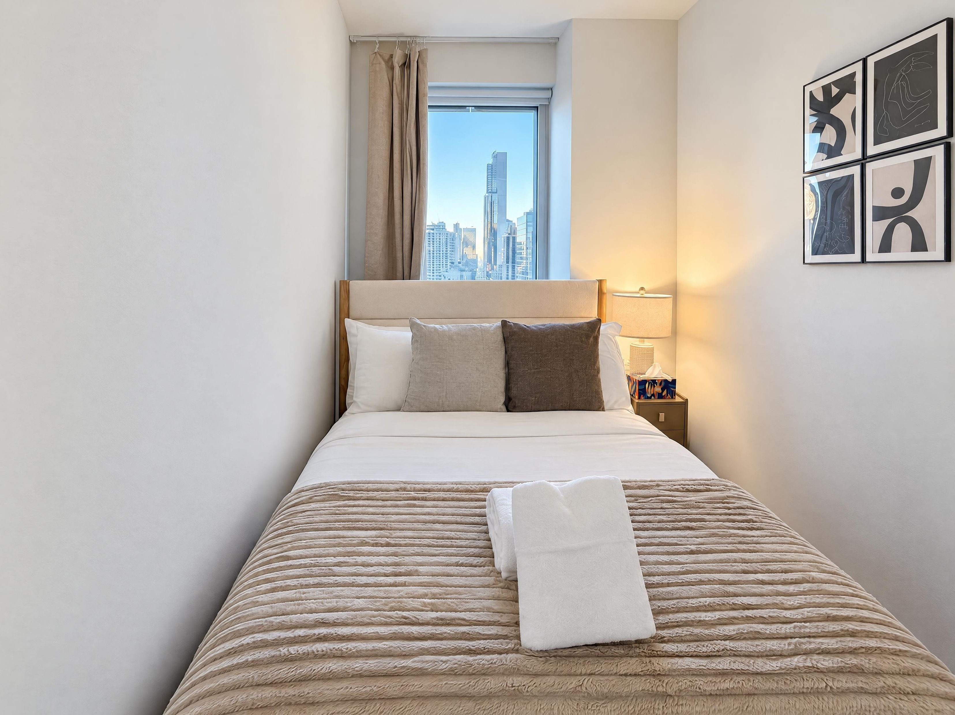 Ein Hotelzimmer mit einem Bett, Kissen, einem Nachttisch mit Lampe und einem Fenster mit Blick auf die Stadt.