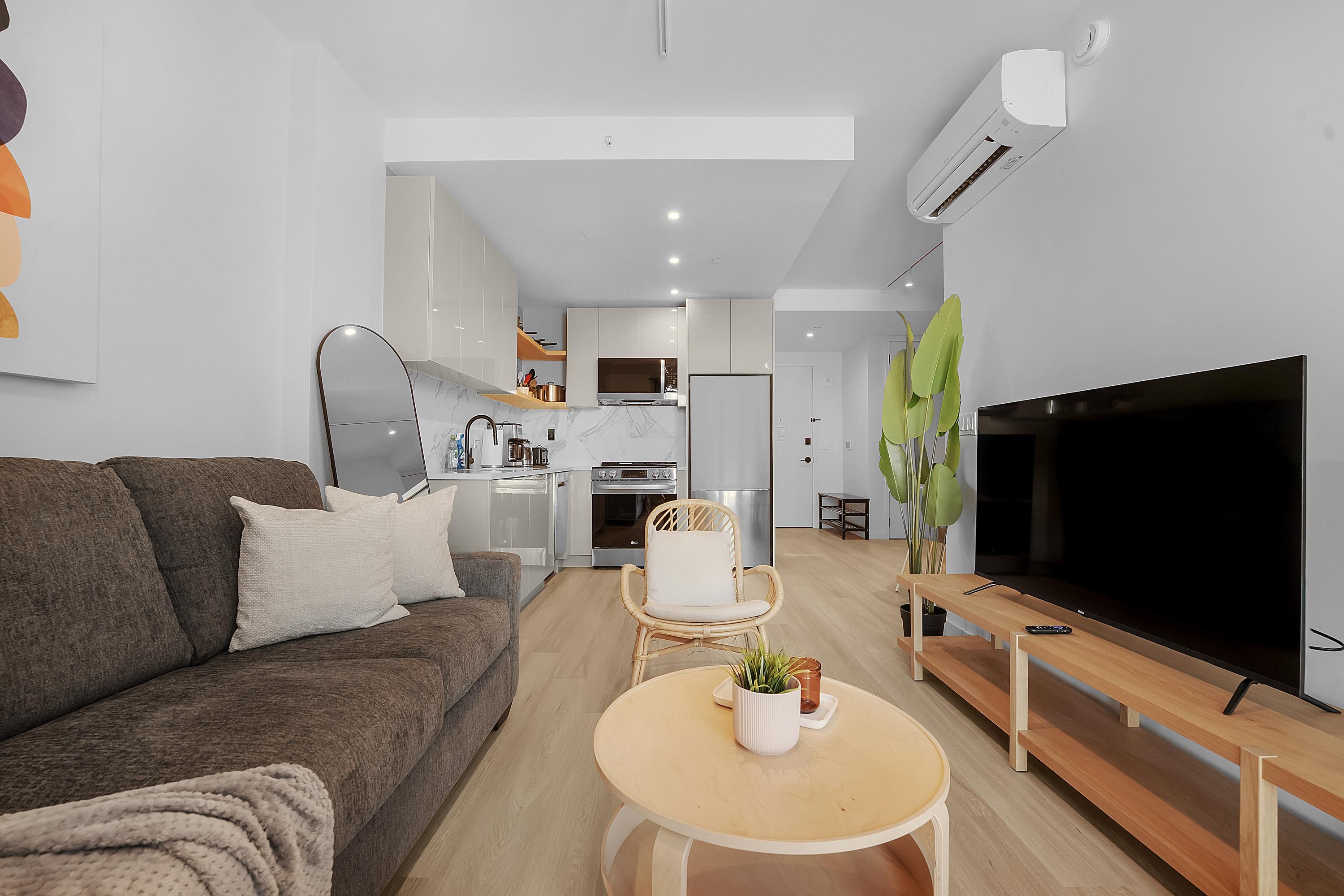 Un salon moderne avec un canapé, une table basse, un téléviseur et une cuisine en arrière-plan.