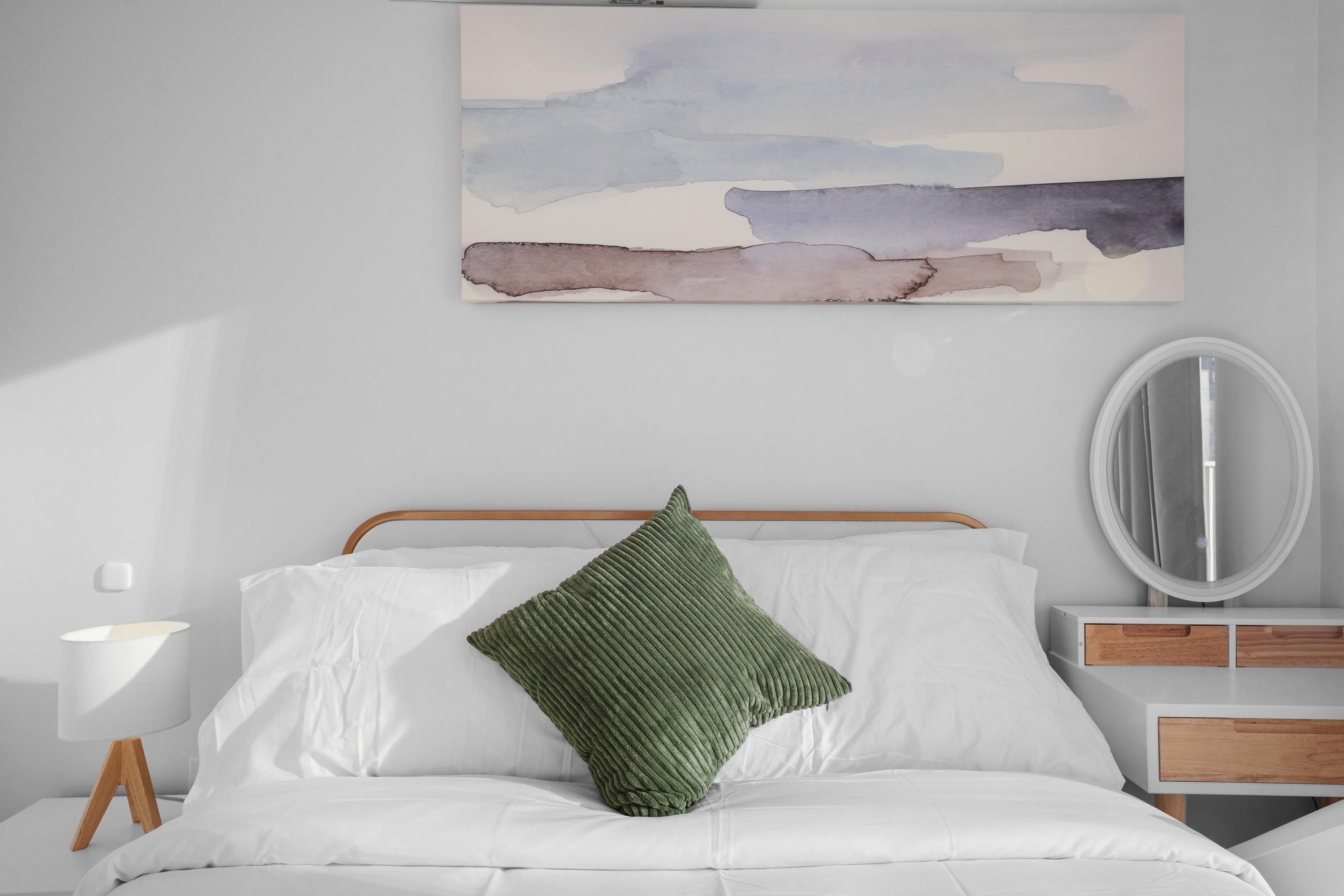 Una cama bien hecha con sábanas blancas y una almohada verde, una mesita de noche con una lámpara, y un espejo montado en la pared.