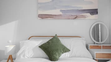 Una cama bien hecha con sábanas blancas y una almohada verde, una mesita de noche con una lámpara, y un espejo montado en la pared.