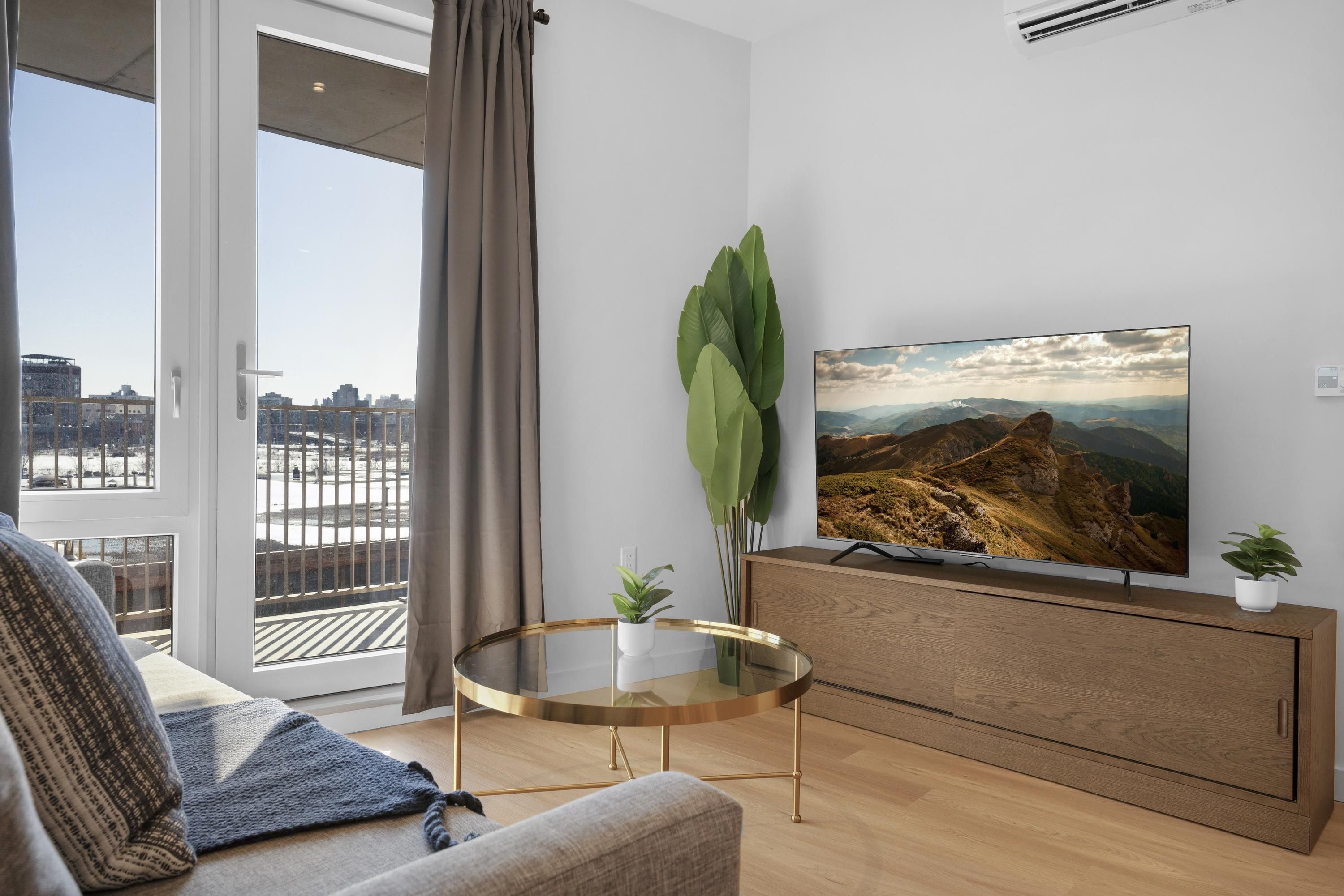 Una sala de estar moderna con un televisor de pantalla plana grande, una mesa de centro de vidrio, un sofá y vistas de un paisaje urbano a través de una puerta corrediza de cristal.
