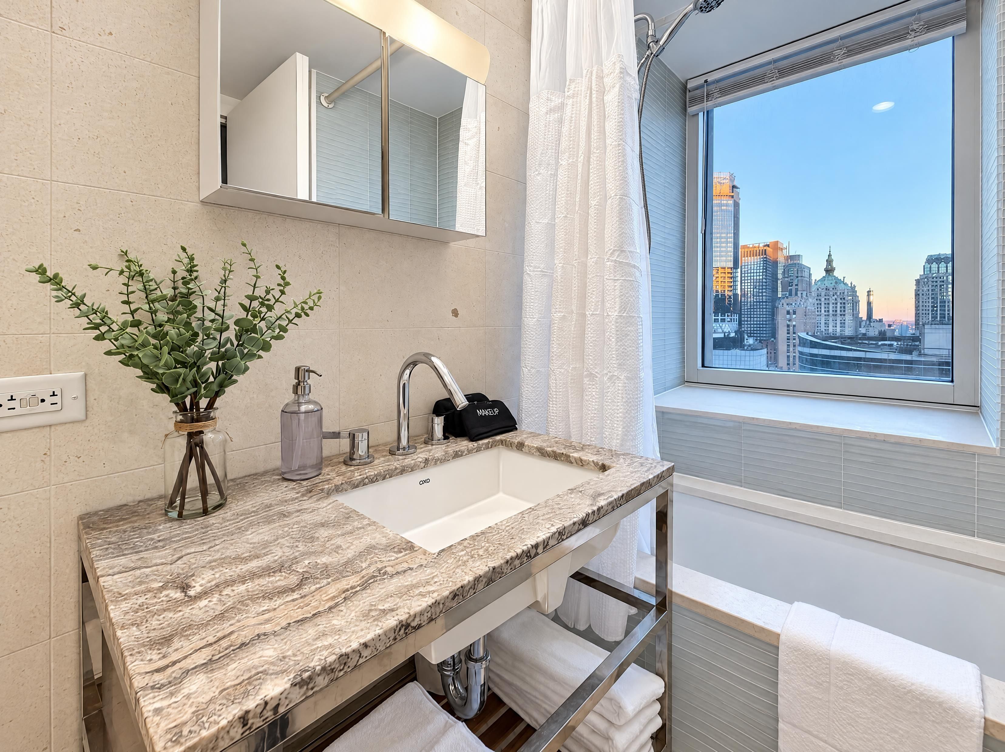 Ein modernes Badezimmer mit Marmorplatte, einem großen Spiegel, einer Handseifenflasche, einer schwarzen Kosmetiktasche und einem Fenster mit Blick auf die Stadt.