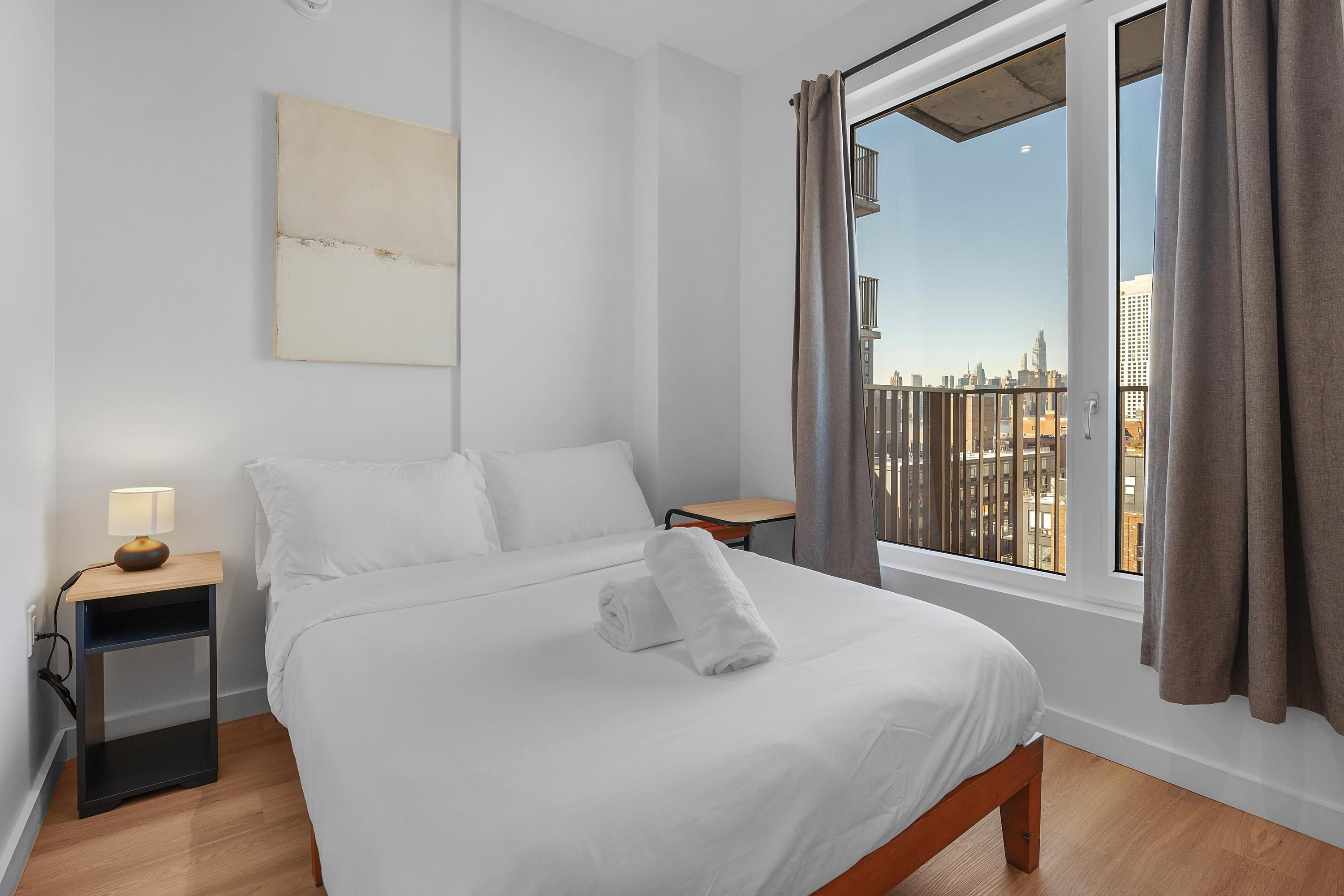 Habitación de hotel con cama, mesita de noche con lámpara, ventana con cortinas y vista a la ciudad.