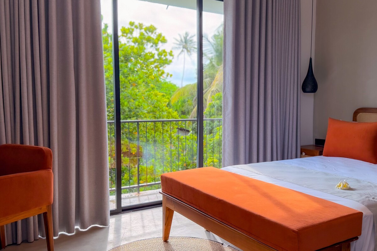 Une chambre d’hôtel avec un lit, une chaise, un balcon donnant sur un espace verdoyant et un luminaire suspendu.