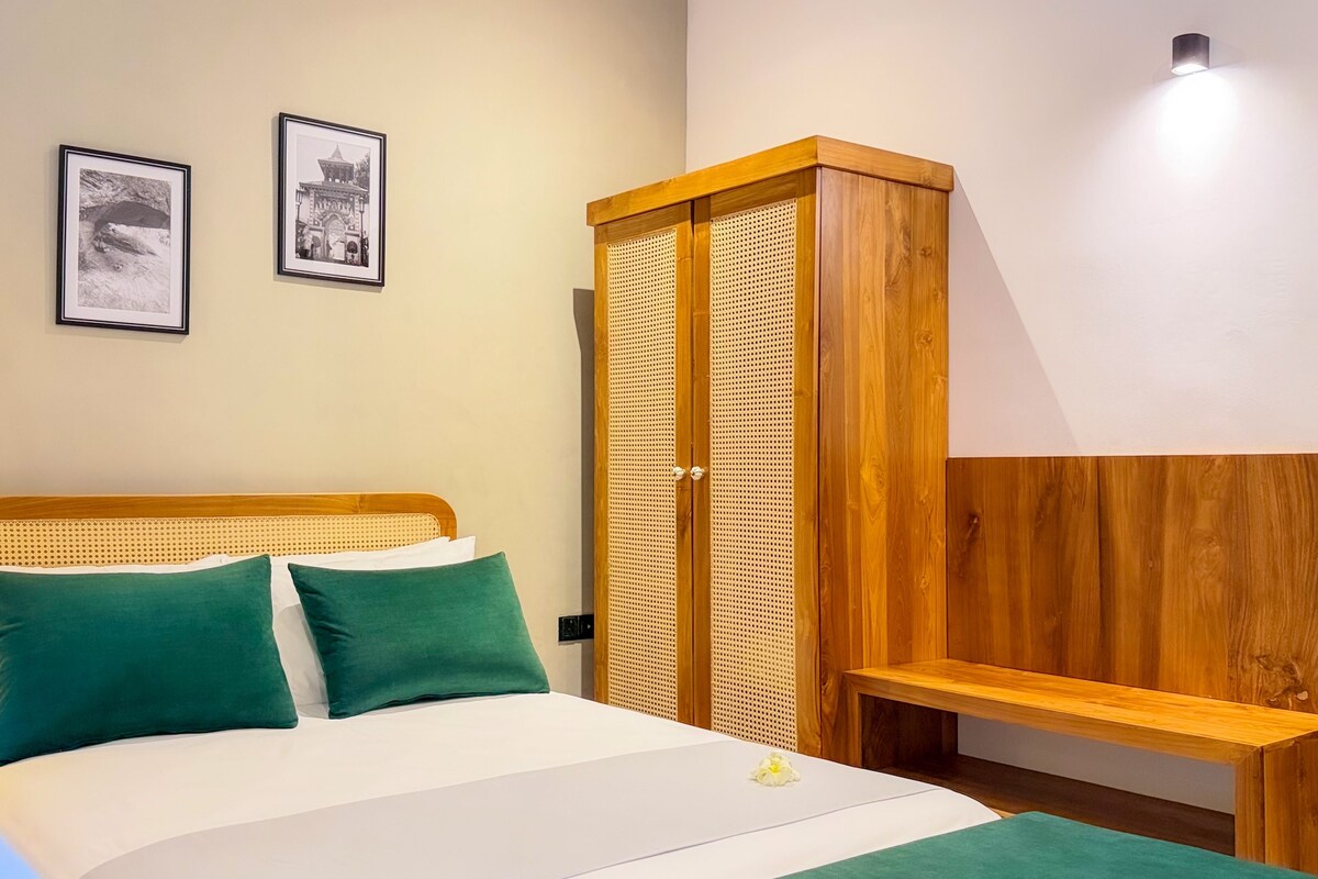 Un lit avec une tête de lit en bois, deux oreillers verts et un couvre-lit blanc. On trouve également dans la pièce un banc en bois et une armoire en osier.