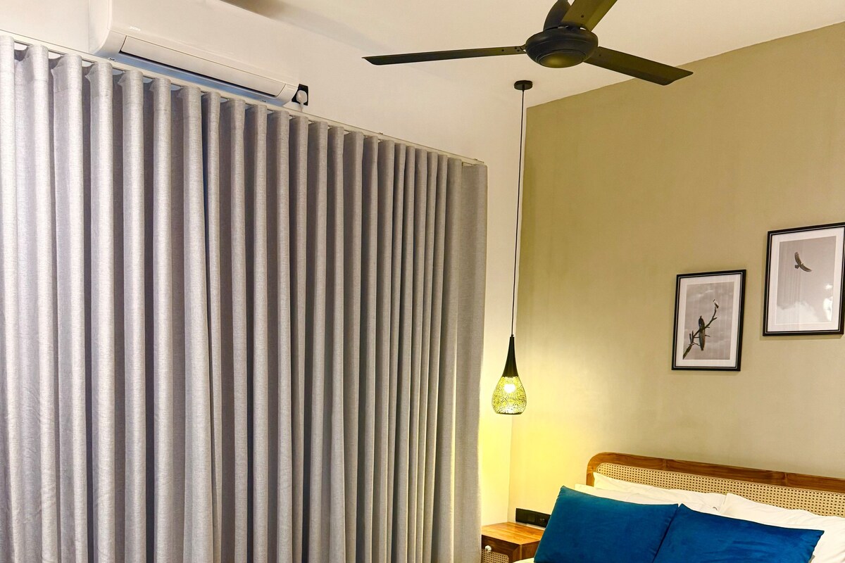 Une chambre à coucher comprenant un lit, un ventilateur de plafond, un appareil de climatisation, une table de chevet avec une lampe et des rideaux.