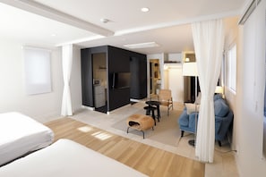 Habitación de hotel moderna con una cama, una zona de estar con mesa y sillas, una kitchenette y un baño visible a través de una puerta abierta.