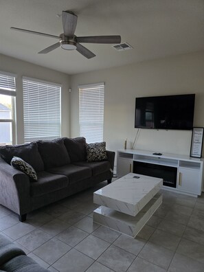 A living room with a ceiling fan, a flat-screen TV mounted on the wall, a dark-colored sofa with patterned cushions, a white coffee table, and a white entertainment unit with a TV. - 3-bedroom house Oasis of calm, in safe neighborhood of Killeen. Airport area. (Killeen)