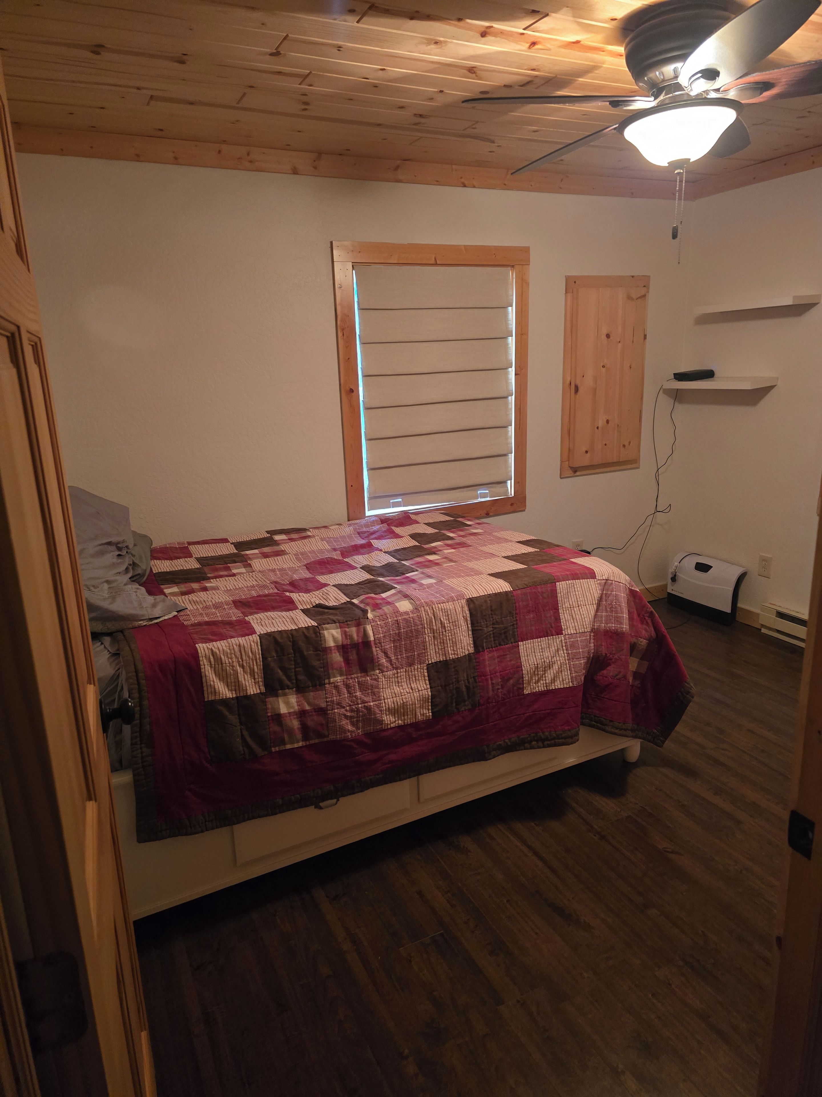 A bedroom with a wooden ceiling, a bed with a quilt, a window with blinds, and a ceiling fan.