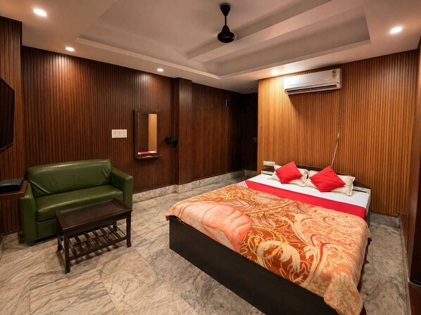 A hotel room with a bed, a green sofa, a small table, wooden paneling, an air conditioning unit, and a ceiling fan.