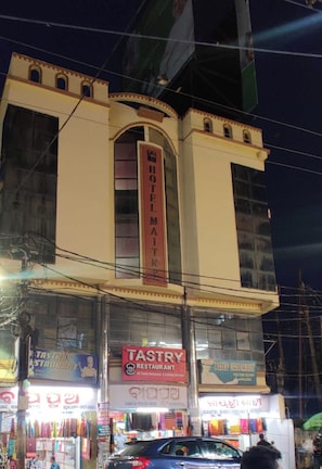 A building with a prominent sign for 'Hotel El Maitre' and multiple storefronts including 'TASTRY RESTAURANT' and 'GAYATRI SAREL'.