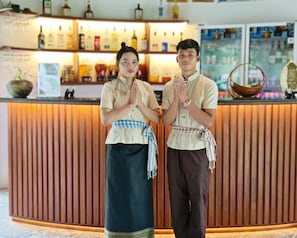 Two individuals standing at a reception desk with a bar area in the background.