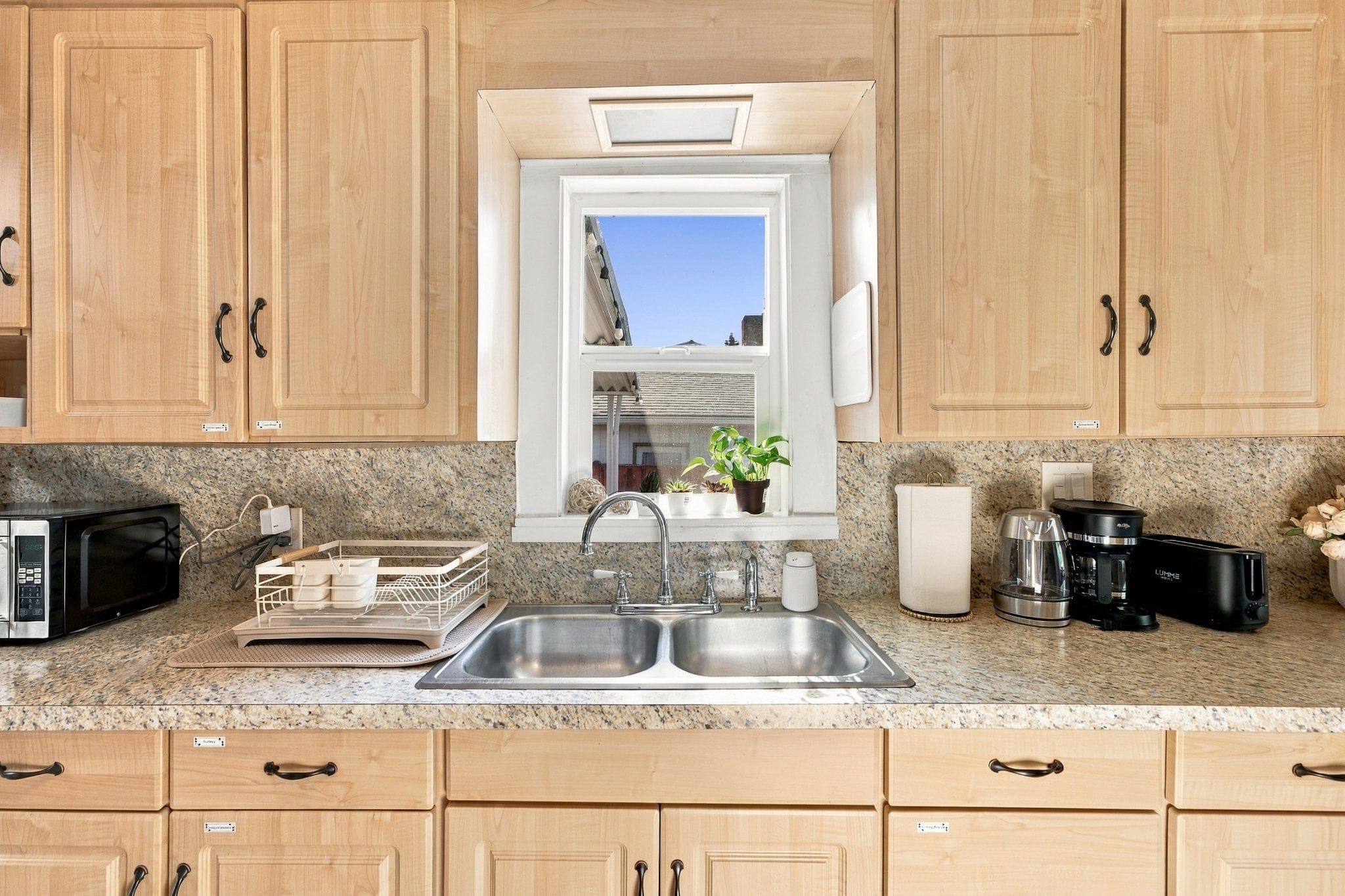 A kitchen with wooden cabinets, a double sink, a microwave, a coffee maker, and a window with a view of the sky.