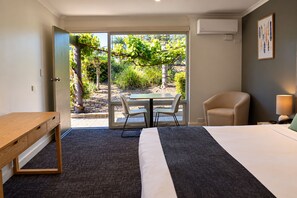 Une chambre d’hôtel comprenant un lit, un bureau, une chaise, une table et donnant sur un jardin grâce à une porte vitrée.