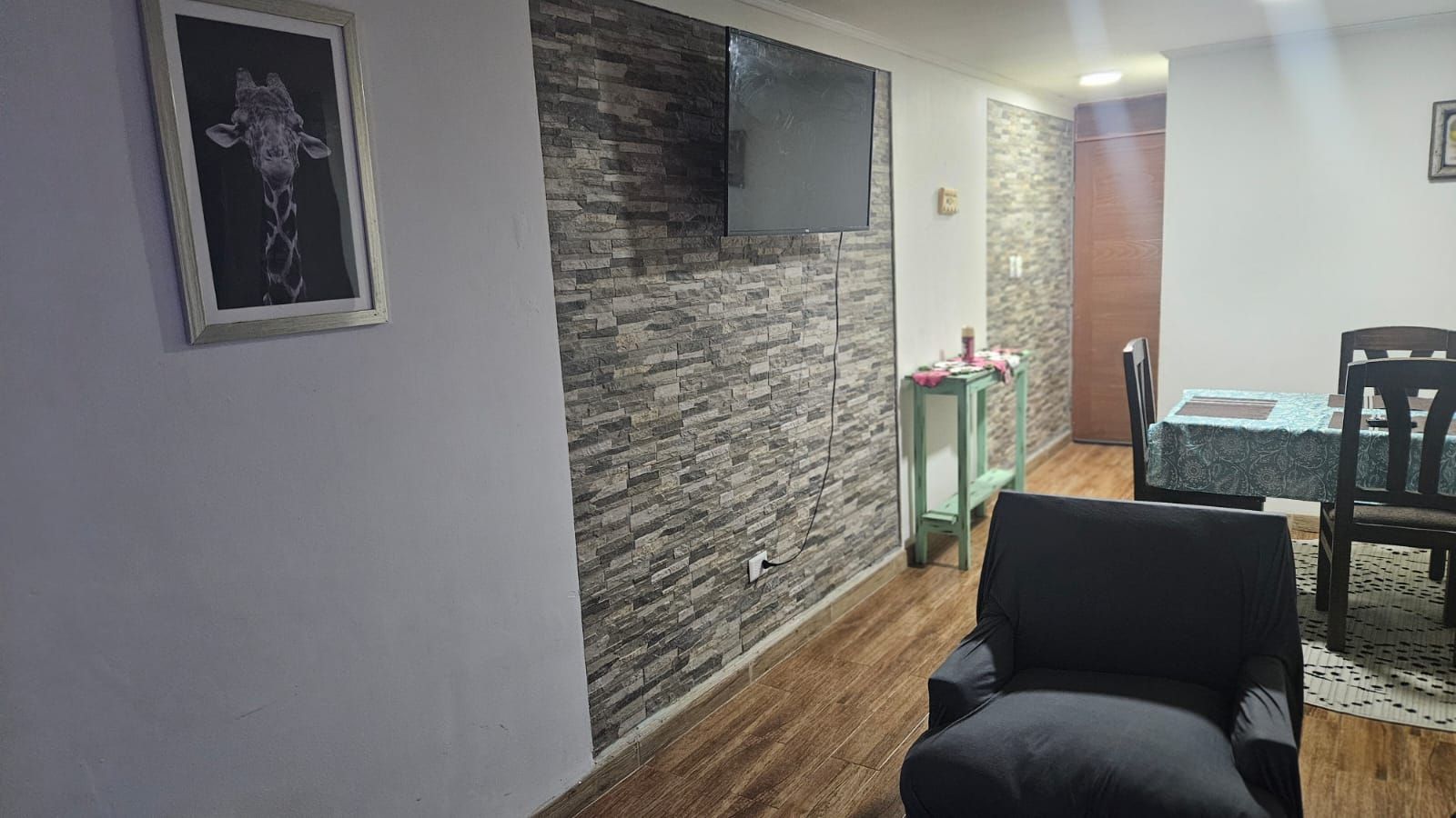 Un salon avec un mur en pierre, un téléviseur fixé au mur, un fauteuil noir et un coin repas comprenant une table et des chaises.