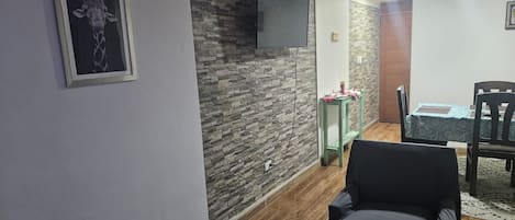 A living room with a stone wall, a mounted TV, a black armchair, and a dining area with a table and chairs.