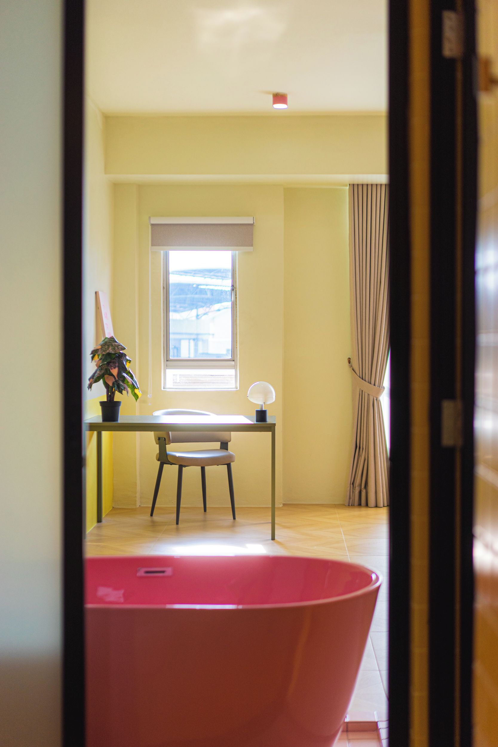 A room with a large bathtub, a window with blinds, a desk with a chair, and a potted plant.