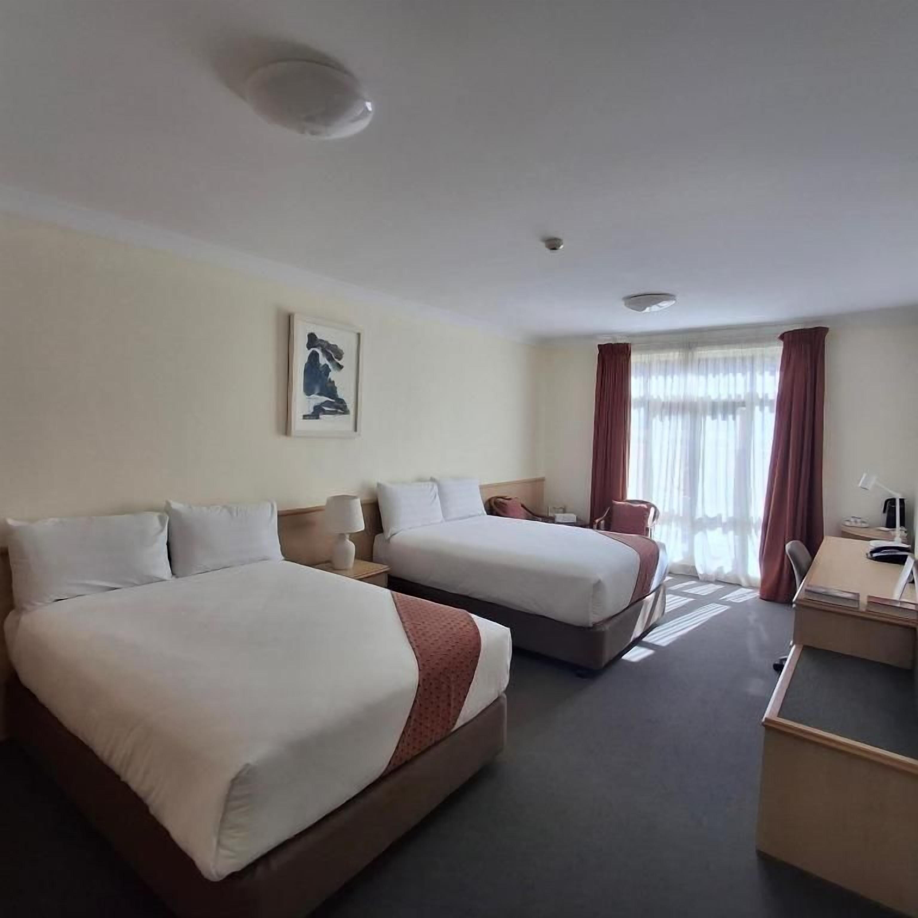 Una habitación de hotel con dos camas, un escritorio, una silla y un ventanal con cortinas.