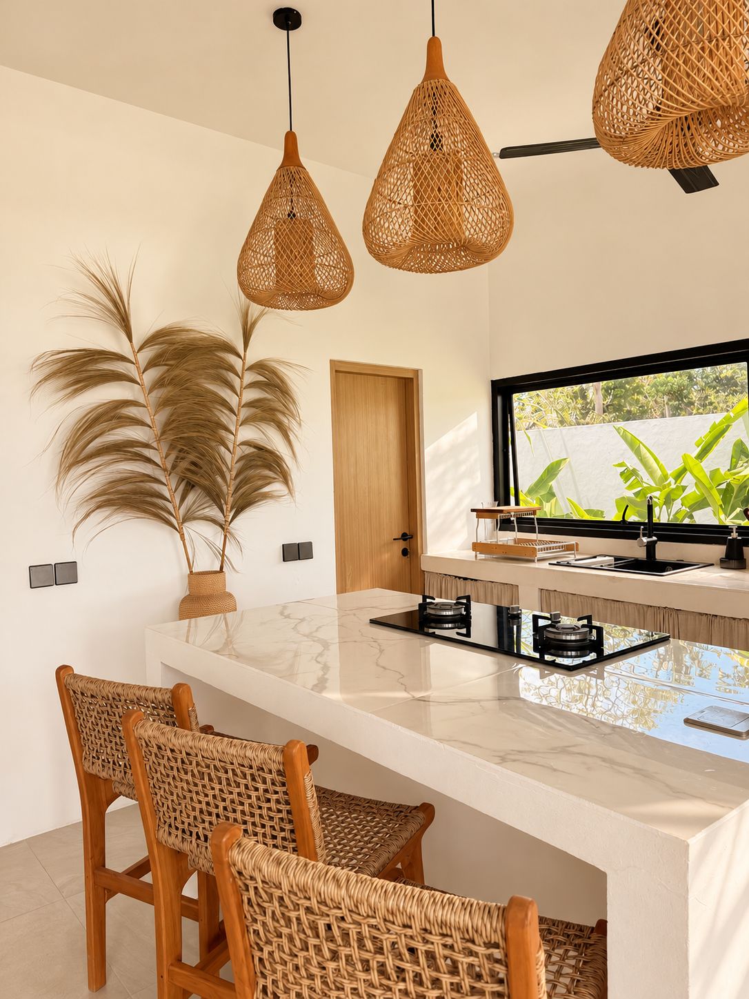 Een moderne keuken met een marmeren aanrechtblad, rieten stoelen en een groot raam met beplanting erbuiten.