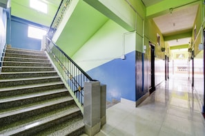 Una escalera con barandilla de metal que lleva a un rellano, un pasillo con una decoración azul y verde, y un suelo de baldosas.