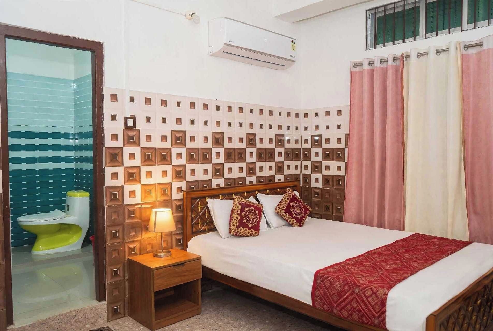 A hotel room with a bed, a bedside table with a lamp, a window with curtains, and a bathroom with a toilet and sink.
