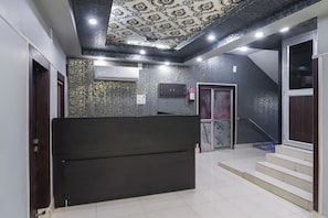 A hotel lobby with a patterned ceiling, a reception desk, a staircase, and a fire extinguisher.