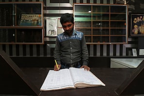 A person sitting at a table with a large open ledger book, writing with a yellow pen. - Hotel O Singh Palace (Darbhanga)