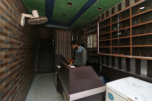 A reception desk with a person seated behind it, a wall-mounted fan, and a window with blinds. - Hotel O Singh Palace (Darbhanga)