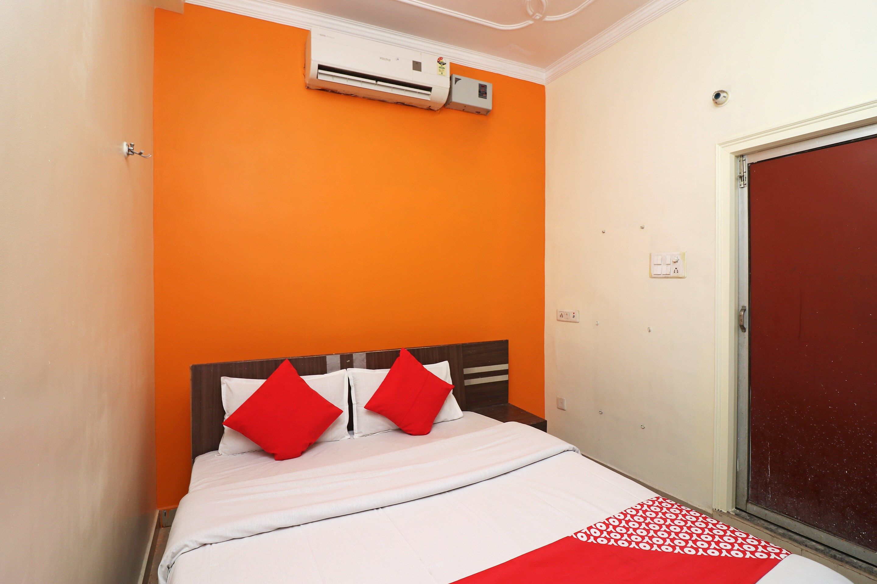 A hotel room with an orange wall, a bed with white bedding and red pillows, an air conditioning unit above the bed, and a red door.