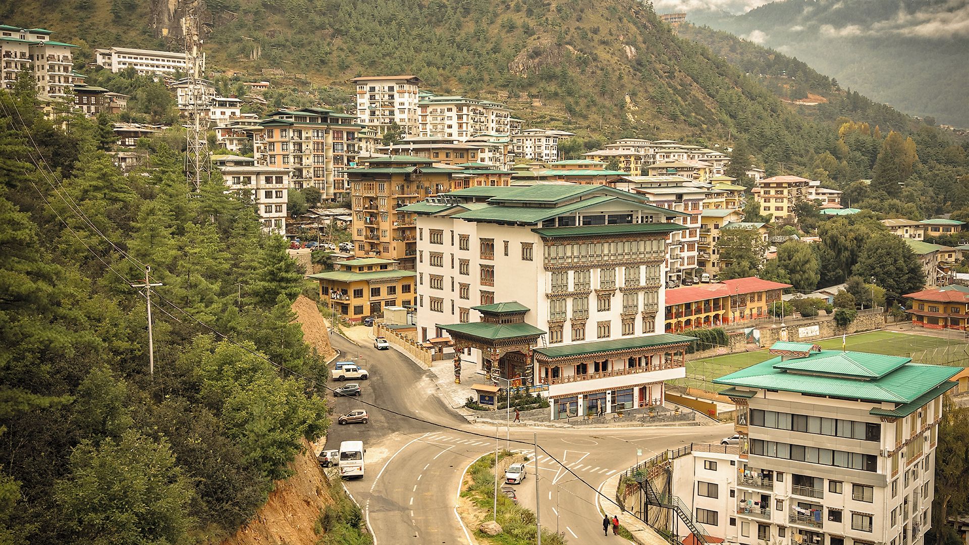A mountainous town with multi-story buildings, a winding road, and lush greenery.