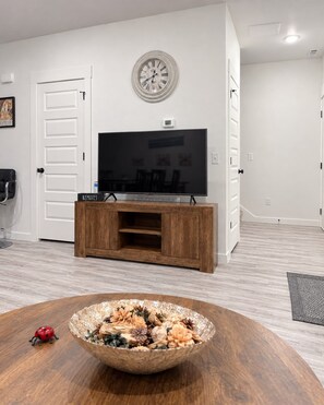 A modern living room with a flat-screen TV on a wooden entertainment unit, a decorative clock on the wall, and a round table with a decorative bowl in the foreground. - Business or Family: 3BR Near Walmart HQ & Trails • Parking (Bentonville)