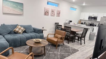 A modern living room with a sofa, coffee table, and chairs, a kitchen area with cabinets and appliances, and artwork on the walls.