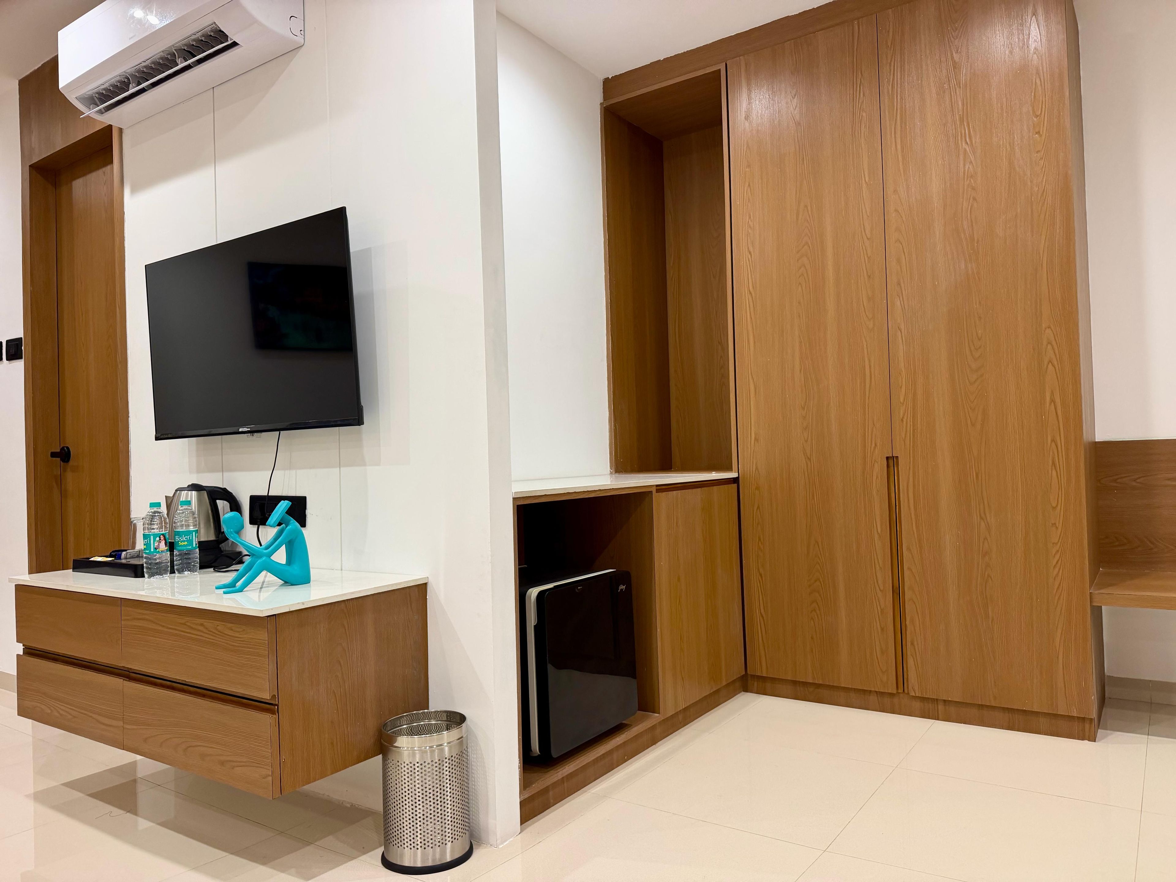 Habitación con televisor de pantalla plana montado en la pared, un mueble de madera, un pequeño cubo de basura y piso de baldosas.