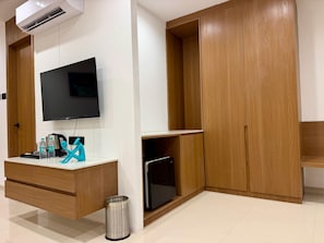 Habitación con televisor de pantalla plana montado en la pared, un mueble de madera, un pequeño cubo de basura y piso de baldosas.
