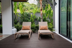Two lounge chairs with cushions on a wooden deck, surrounded by lush greenery and a modern building facade. - Quiet 3BR Pool Villa in Soi Naya near Nai Harn Beach (Tambon Rawai)