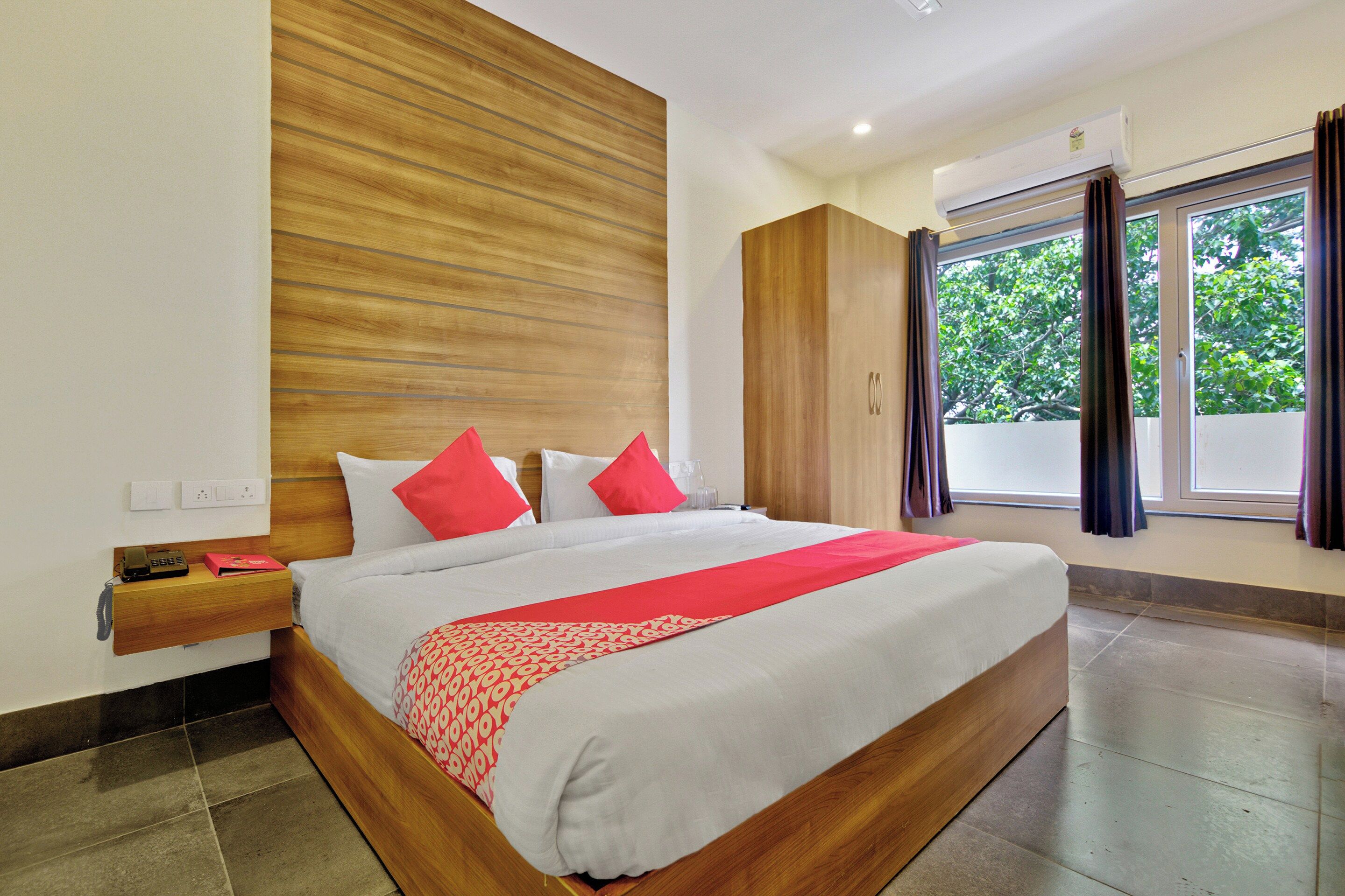 Una habitación de hotel con cabecera de madera, una cama grande con ropa de cama blanca y roja, una mesita de noche con teléfono y una ventana con cortinas.