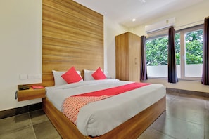 Una habitación de hotel con cabecera de madera, una cama grande con ropa de cama blanca y roja, una mesita de noche con teléfono y una ventana con cortinas.