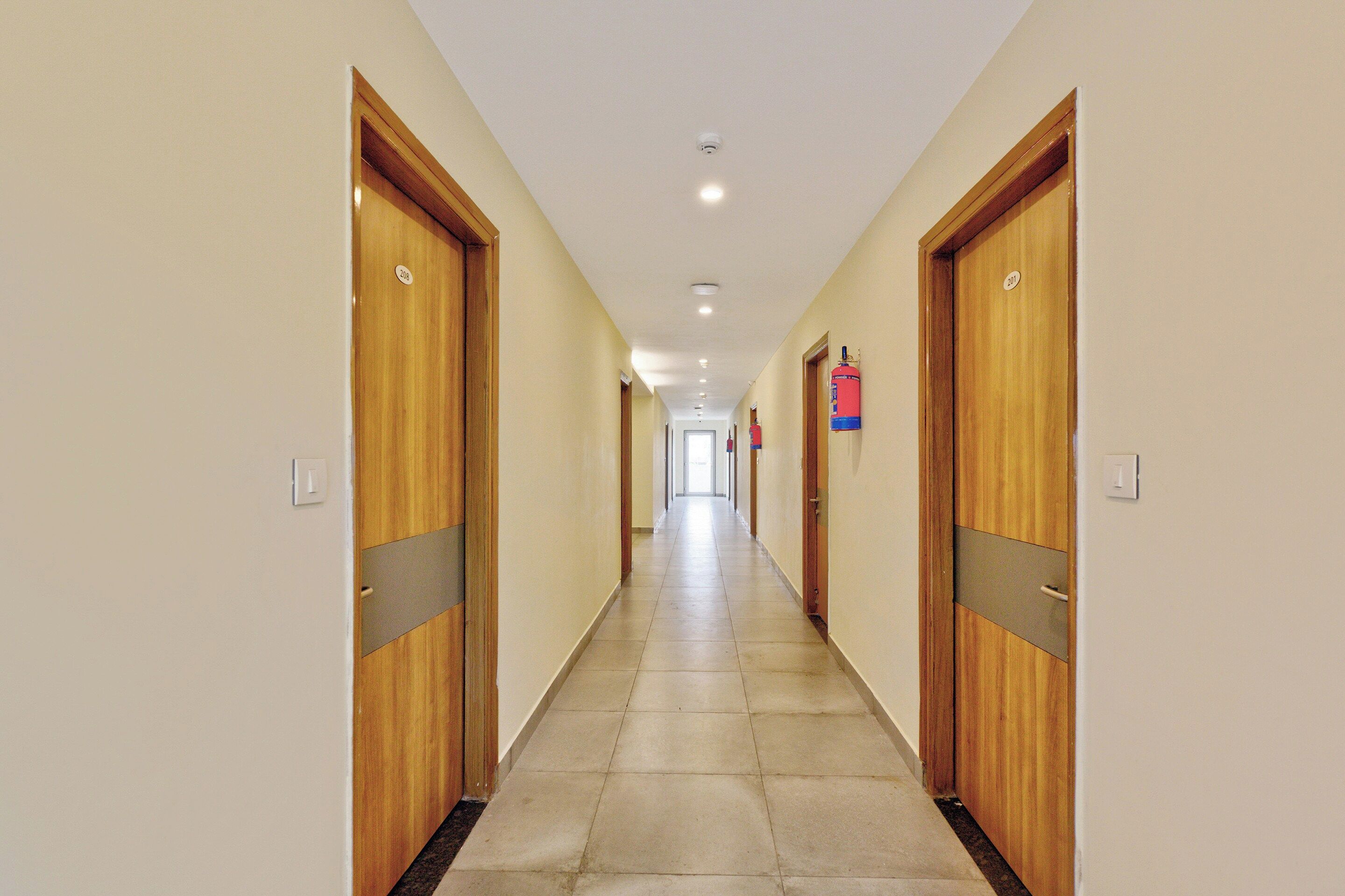 Un pasillo con puertas de madera, placas con números y un extintor.