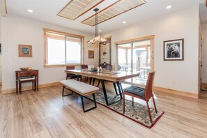 A dining area with a wooden table, bench, and chairs, a hanging light fixture, and a view through large windows.