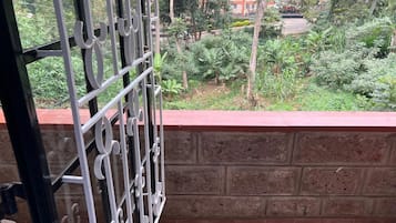 Ein Balkon mit einem Metallgitter, einer Ziegelwand und Blick auf Grünflächen und Gebäude im Hintergrund.