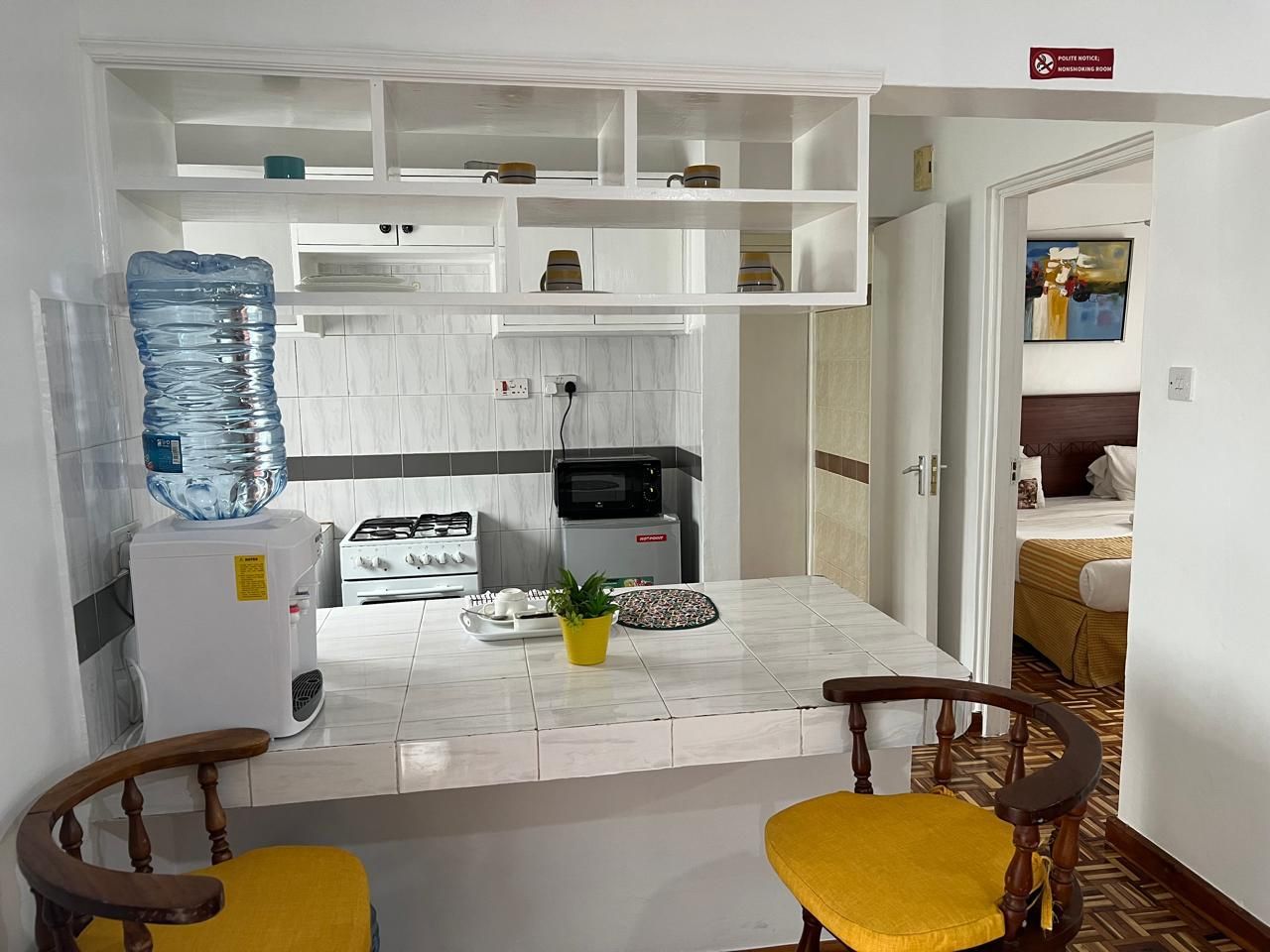 Une cuisine compacte avec des meubles blancs, un distributeur d’eau, une cuisinière et un four à micro-ondes. On y trouve deux chaises en bois avec des coussins jaunes.