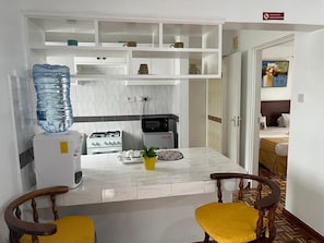 Une cuisine compacte avec des meubles blancs, un distributeur d’eau, une cuisinière et un four à micro-ondes. On y trouve deux chaises en bois avec des coussins jaunes.