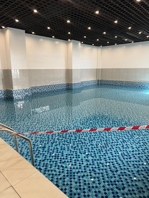 Une piscine intérieure dont le sol est recouvert de carreaux disposés en motif à damier, avec une bande de signalisation jaune le long du bord et une échelle en métal.