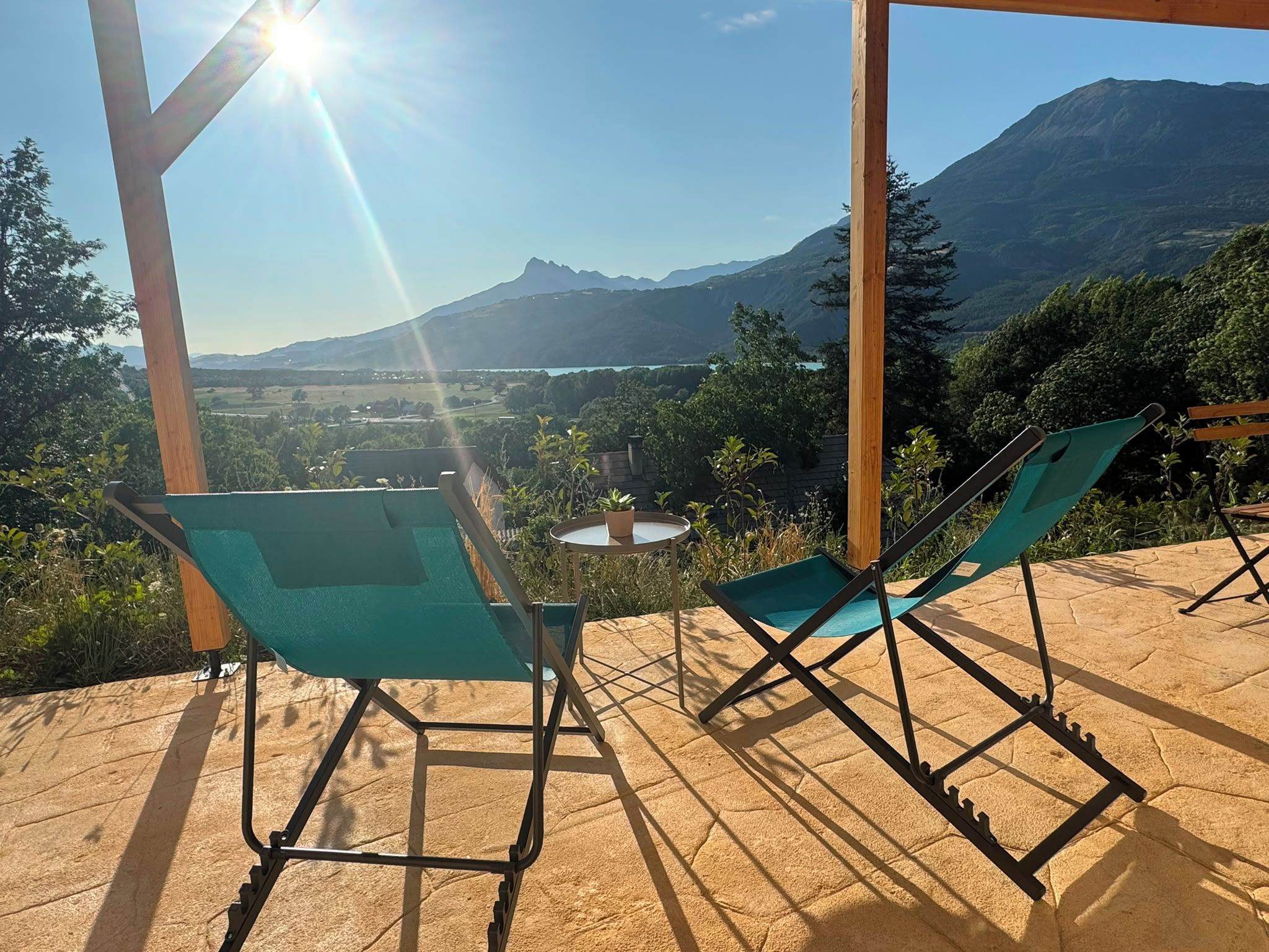 A terrace with two green chairs and a small round table, overlooking a mountainous landscape.