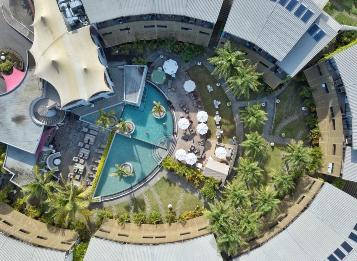 Luchtbeeld van een hotel met een rond zwembad, omringd door palmbomen en buiten zitgedeeltes.